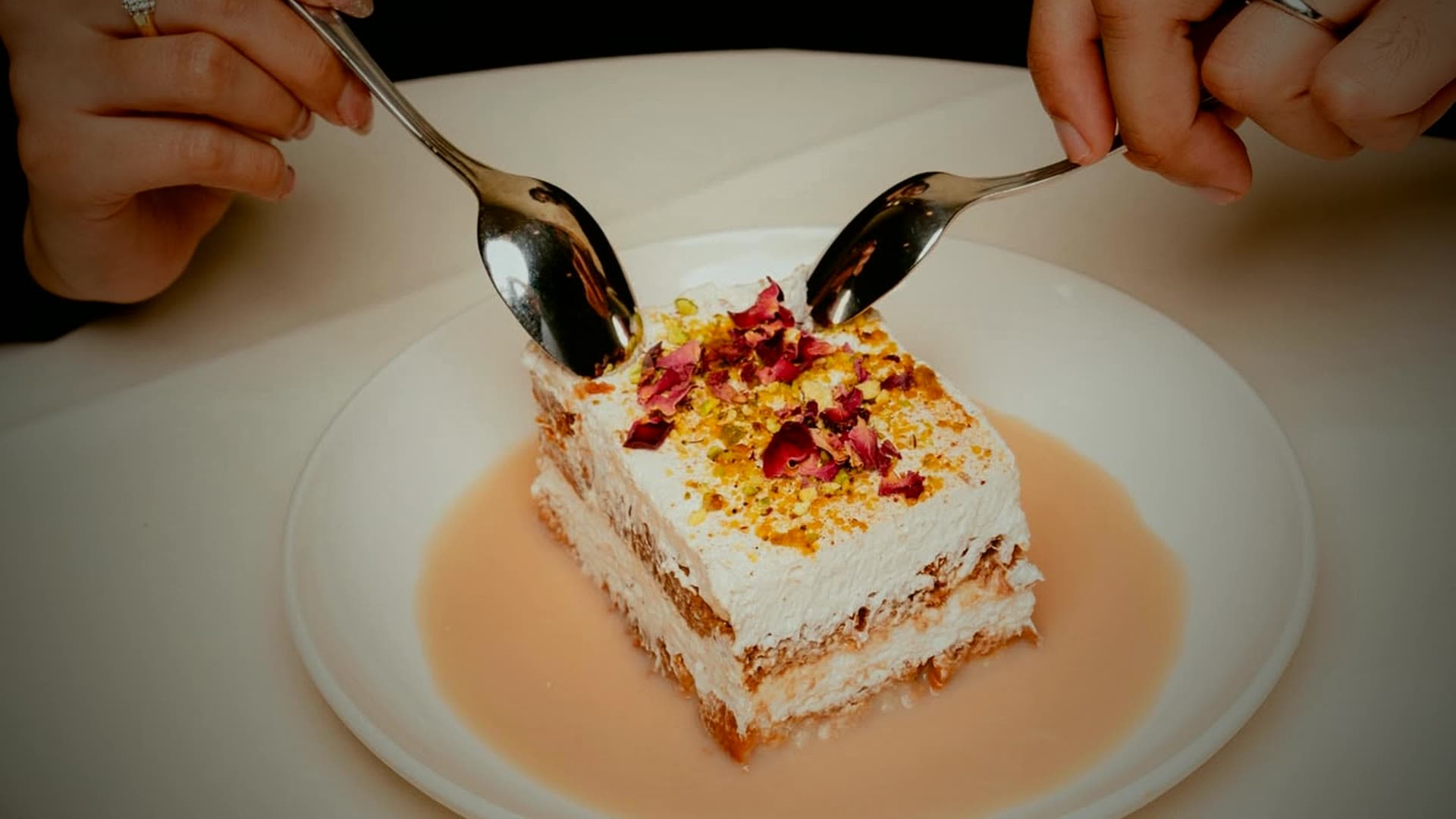 A plated rose-and-pistachio tres leches dessert, two spoons reaching in